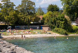 Mensen genieten van de zon en zwemmen aan een meerstrand nabij Divonne Les Bains, omringd door groen.