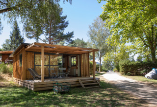 Wooden cabin with covered terrace in a sunny, green holiday park at Huttopia Lac de la Siauve, France.