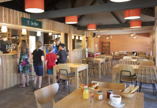 Binnenzicht van de cafetaria in Huttopia Lac de la Siauve met klanten aan de toog en ontbijt op tafels.
