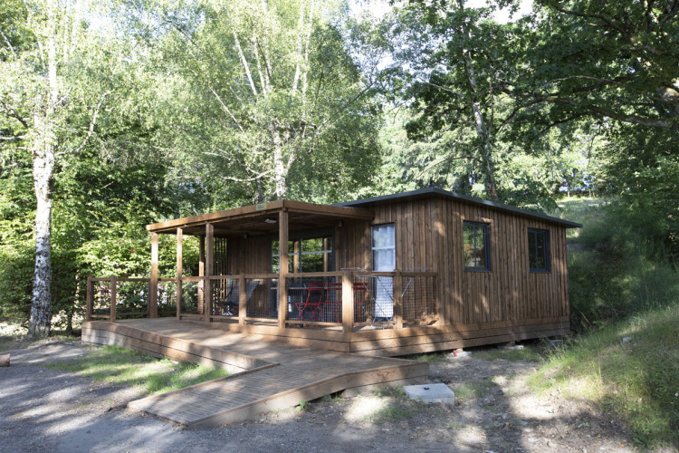 Wooden cabin with a covered porch surrounded by lush trees at Huttopia Lac de la Siauve holiday park, France.