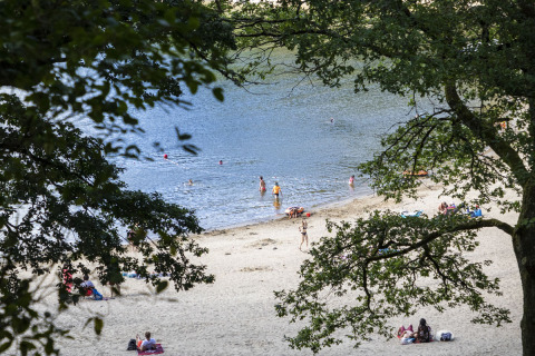 Vista de la playa en Huttopia Lac de la Siauve, con familias relajándose y nadando en el lago al fondo.