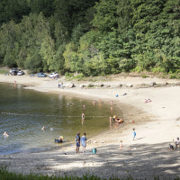 Personas disfrutan de un día soleado en la playa de un lago cerca de Lanobre, rodeada de bosques verdes.