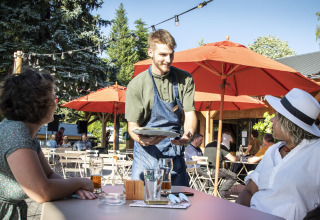 Camarero atiende a dos mujeres en cafetería al aire libre en Huttopia Lac de la Siauve, Francia.