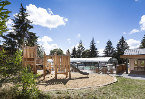 Parque infantil de madera con tobogán junto a una piscina cubierta en un entorno vacacional natural.