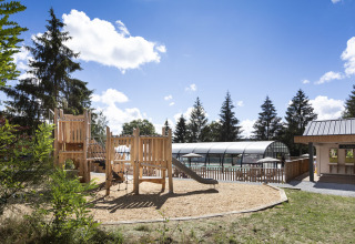 Spielplatz mit Holzturm und Rutsche neben überdachtem Pool in einem bewaldeten Ferienresort.