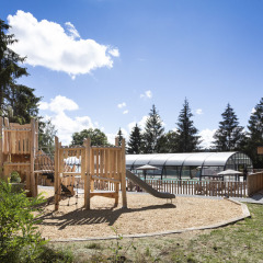 Parque infantil de madera con tobogán junto a una piscina cubierta en un entorno vacacional natural.