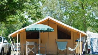 Tienda safari Toile&bois Classic con sillas, mesa y sombrilla en una plataforma de madera rodeada de árboles.