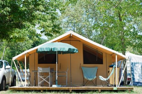 Tienda safari Toile&bois Classic con sillas, mesa y sombrilla en una plataforma de madera rodeada de árboles.