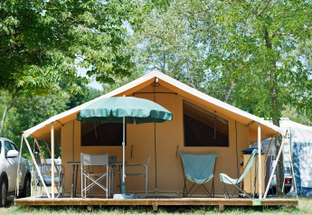 Tenda safari Toile&bois Classic con sedie, tavolo e ombrellone su pedana in legno tra alberi verdi.