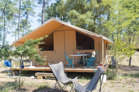 Tienda safari Toile&bois Classic en un camping forestal con terraza de madera y área de asientos al aire libre.