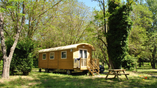 Cabaña de madera estilo caravana gitana en claro del bosque, con mesa de picnic y coche detrás.