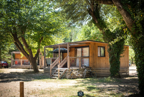 Cabaña de madera con porche y bicicletas delante, entre árboles, en Huttopia le Moulin, Francia.