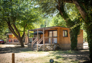 Cabaña de madera con porche y bicicletas delante, entre árboles, en Huttopia le Moulin, Francia.