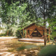 Safari-teltet Tent Saadani midt i en skovlysning med terrasse og bænk, omgivet af grønne træer.