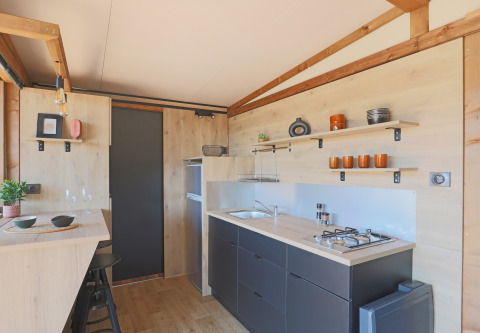 Cocina de tienda safari moderna con detalles de madera, estantes abiertos y comedor en Huttopia Vallée du Lot, Francia.
