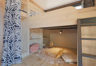 Zona de litera acogedora con cortinas estampadas y rincón de lectura en una tienda safari de Huttopia Vallée du Lot, Francia.