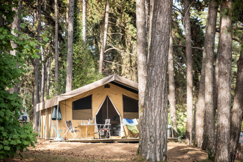 Tienda de safari Toile&Bois Sweet en un bosque de pinos, con terraza, sillas y mesa para camping.