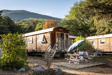 To personer ved picnicbord foran træhytte 'Gypsy caravan' i skovklædt bjergområde om sommeren.