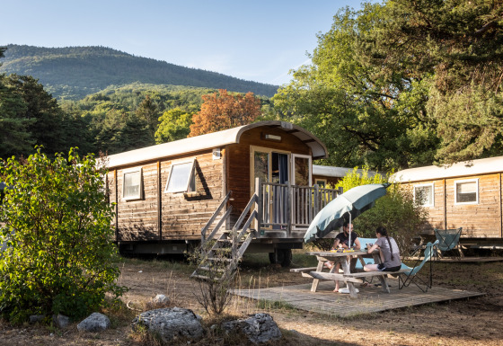 To personer ved picnicbord foran træhytte 'Gypsy caravan' i skovklædt bjergområde om sommeren.