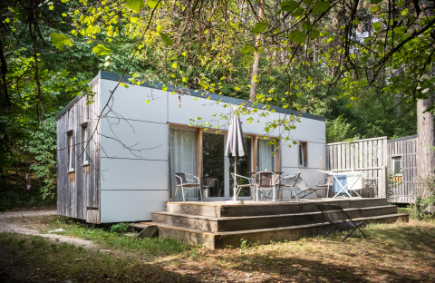 Casa móvil Vancouver en Huttopia Divonne Les Bains, Francia, cabaña rodeada de árboles y terraza de madera.