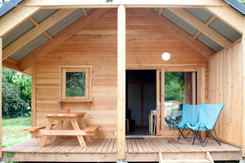 Cabaña de madera con porche, mesa de picnic y dos sillas azules en Huttopia Divonne Les Bains, Francia.