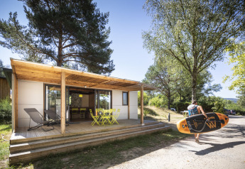 En person bærer et paddleboard foran Mobile home Vancouver-cabin med terrasse og havemøbler.
