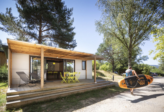 En person bærer et paddleboard foran Mobile home Vancouver-cabin med terrasse og havemøbler.