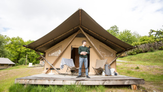Hombre de pie frente a una tienda safari Trapper con terraza de madera y sillas de camping al aire libre.