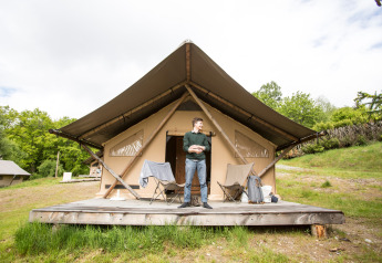 Uomo in piedi davanti a una tenda safari Trapper con pedana in legno, sedie e natura circostante.