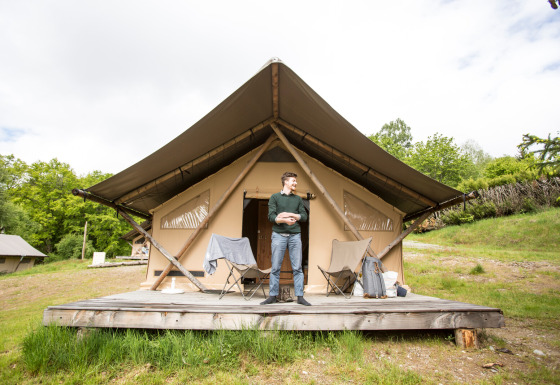 Mann steht vor einem Trapper Safari-Zelt auf einer Holzterrasse, umgeben von Natur und Stühlen.