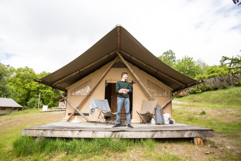 Homme debout devant une tente safari Trapper avec terrasse en bois, chaises de camping et nature autour.