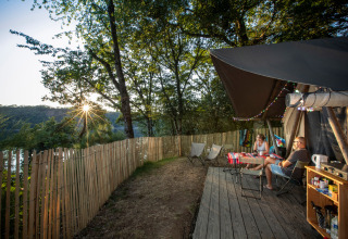 Two people relax outside a Trapper safari tent at Huttopia Lac de la Siauve in France, surrounded by trees and sunlight.