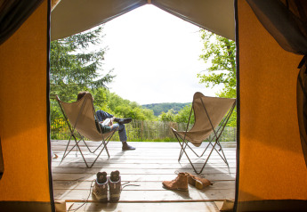 Blick aus einem Safarizelt auf eine Holzterrasse mit Stühlen und Schuhen bei Huttopia Lac de la Siauve.