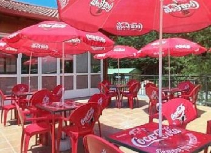 Buiten zitgedeelte bij Camping Etxarri in Navarra, Spanje, met rode Coca-Cola stoelen, tafels en parasols.