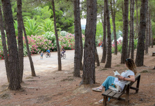 Woman reading on bench among pine trees while kids cycle nearby at TAIGA Lake Caspe, Aragón, Spain.
