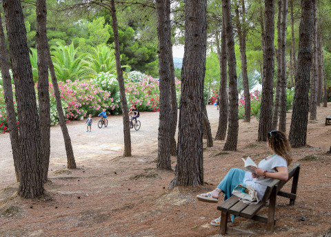 Frau liest auf einer Bank im Wald, während Kinder in der Nähe bei TAIGA Lake Caspe, Aragón, Spanien Rad fahren.