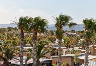 Palmen und moderne Hütten in L'Ampolla, Katalonien, Spanien, mit Bergen im Hintergrund an einem sonnigen Tag.