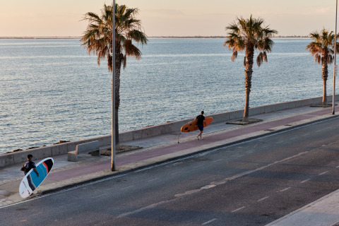 Two surfers walk along a seaside promenade with palm trees near L'Ampolla, Catalonia, Spain at sunset.