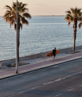 Dos surfistas caminan por el paseo marítimo con palmeras cerca de L'Ampolla, Cataluña, España al atardecer.
