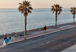 Dos surfistas caminan por el paseo marítimo con palmeras cerca de L'Ampolla, Cataluña, España al atardecer.