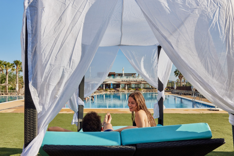 Twee mensen ontspannen onder een cabana aan het zwembad in TAIGA Delta de l'Ebre vakantiepark, Catalonië, Spanje.
