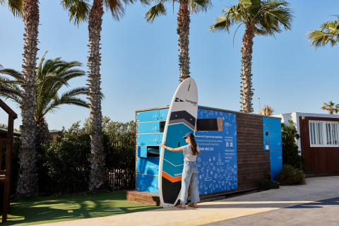 Vrouw haalt een paddleboard uit een lockersysteem bij TAIGA Delta de l'Ebre, Catalonië, tussen palmbomen.