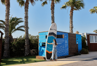 Vrouw haalt een paddleboard uit een verhuurkluis bij TAIGA Delta de l'Ebre, Catalonië, omringd door palmbomen.