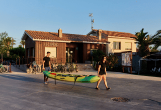 Zwei Personen tragen ein Kajak vor der Rezeption im TAIGA Delta de l'Ebre Ferienpark in Katalonien, Spanien.