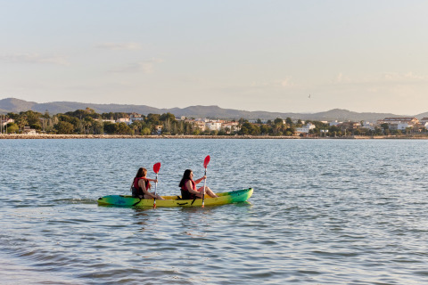 Dos personas en kayak reman por el agua cerca de L'Ampolla, Cataluña, con la ciudad y colinas al fondo.