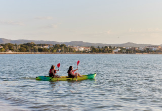Two people kayaking on the water near L'Ampolla, Catalonia, Spain, with the town and hills in the background.