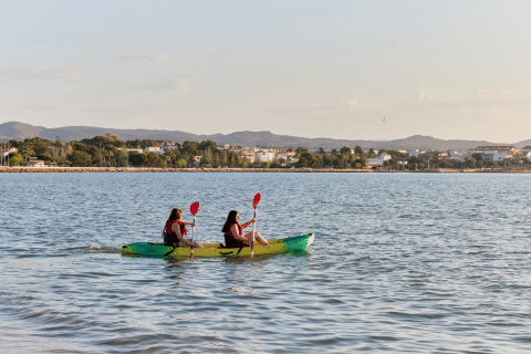 Zwei Personen paddeln im Kajak auf dem Wasser bei L'Ampolla, Katalonien, mit Stadt und Bergen im Hintergrund.