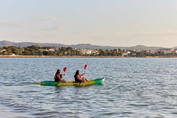 Two people kayaking on the water near L'Ampolla, Catalonia, Spain, with the town and hills in the background.