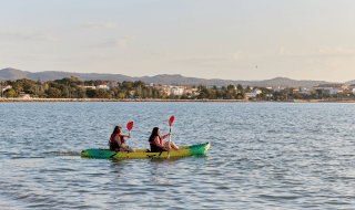 Dos personas en kayak reman por el agua cerca de L'Ampolla, Cataluña, con la ciudad y colinas al fondo.