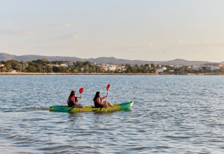 Due persone fanno kayak sull’acqua vicino a L’Ampolla, Catalogna, con la città e le colline sullo sfondo.